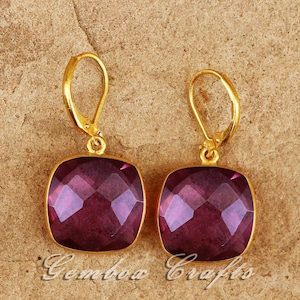 Puede incluir: Un par de pendientes dorados con cierre de palanca y piedras preciosas cuadradas, facetadas, de color púrpura intenso. Los pendientes están sobre un fondo texturizado y beige. El texto "Gembox Crafts" es visible.