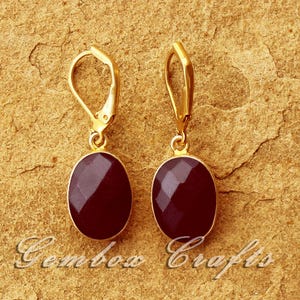 Op de afbeelding: Een paar goudkleurige oorhangers met een ovale, gefacetteerde, donkerrode edelsteen. De oorbellen worden gepresenteerd op een getextureerd, bruin oppervlak. De tekst "Gembow Crafts" staat onderaan de afbeelding.
