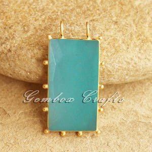 Può includere: Un ciondolo rettangolare con una pietra color turchese incastonata in una cornice dorata. La cornice ha piccoli accenti dorati rotondi lungo i bordi e due piccoli anelli nella parte superiore. Il testo "Gembow Crafts" è visibile.
