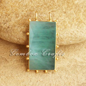 Puede incluir: Un colgante rectangular con un marco dorado y una piedra de color verde azulado. La piedra tiene un aspecto veteado. El marco está adornado con pequeñas cuentas doradas. El texto "Gembow Crafts" es visible en el colgante.