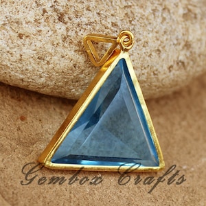May include: A gold-toned pendant featuring a triangular blue gemstone. The pendant is on a light brown surface.