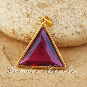 May include: A gold-toned pendant featuring a triangular-shaped red gemstone. The pendant is on a brown surface.
