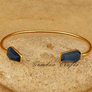 May include: A gold bangle bracelet with two dark blue faceted gemstone accents. The bracelet is open and rests on a light brown stone.