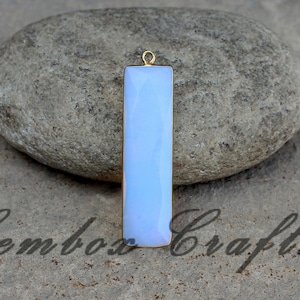 May include: A rectangular, pale blue gemstone pendant with a gold-colored frame and a small loop at the top. The pendant is set against a neutral stone background. The text "Gembos Crafts" is visible in the image.