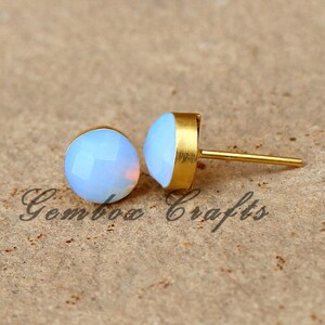 May include: A pair of gold stud earrings with white opal stones. The earrings are on a brown surface.