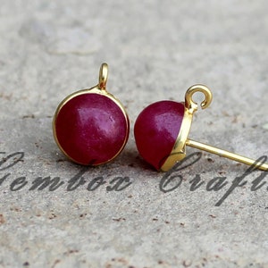 Op de afbeelding: Een paar gouden oorbellen met een ronde, rode edelsteen cabochon. De oorbellen hebben een eenvoudig, elegant ontwerp en zijn perfect voor dagelijks gebruik.