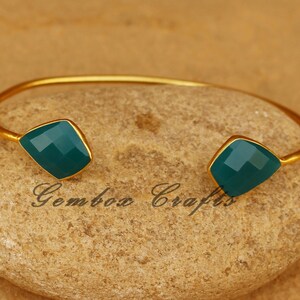 May include: Gold bangle bracelet with two green gemstone accents. The bracelet is open and adjustable.