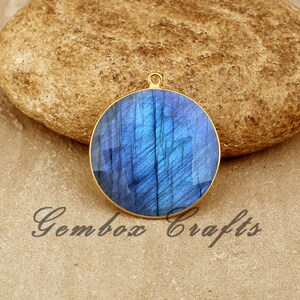 Puede incluir: Un colgante redondo de color dorado con una piedra preciosa de labradorita azul. La piedra preciosa tiene un efecto brillante e iridiscente.