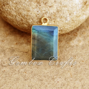 Pode incluir: Um pingente dourado com uma pedra de labradorita retangular. A pedra preciosa é de cor azul escuro, verde e cinza com flashes iridescentes.