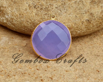 May include: A round, lavender gemstone pendant with a gold-toned bezel setting. The pendant is on a brown, textured surface.