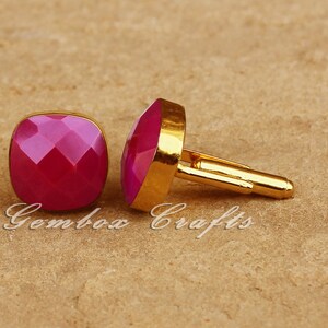 May include: A pair of gold-toned cufflinks with faceted, square, magenta-colored stones. The cufflinks are displayed on a textured, tan surface. The text "Gembox Crafts" is visible in the image.