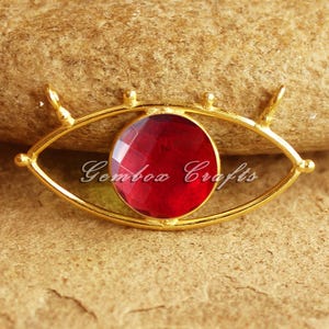 May include: A gold-colored eye-shaped pendant with a large, faceted, round, red gemstone center. The pendant has small loops at the top and bottom for attaching to a chain. The text "Gembox Crafts" is printed below the gemstone.