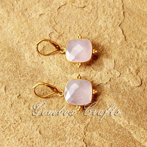 May include: A pair of gold earrings featuring pink square-shaped gemstones. The earrings have a delicate design with small gold beads around the edges of the gemstones.