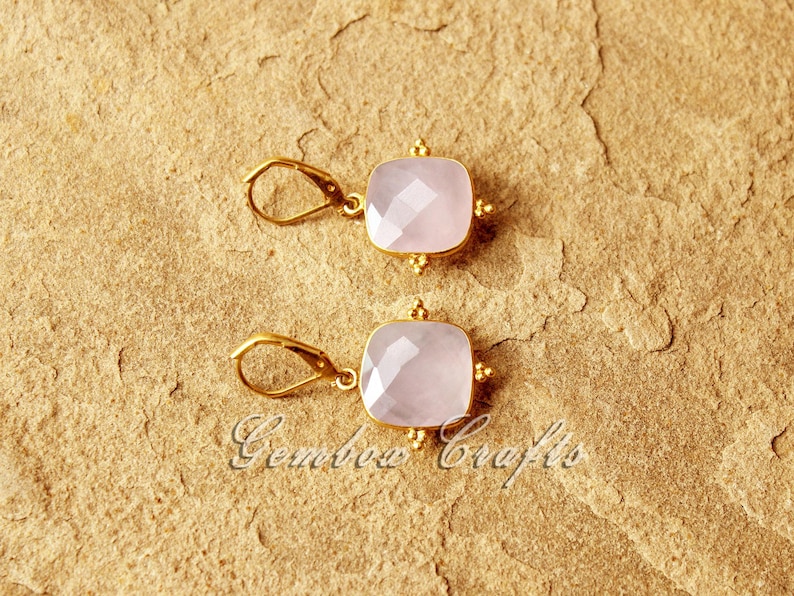 May include: A pair of gold earrings featuring pink square-shaped gemstones. The earrings have a delicate design with small gold beads around the edges of the gemstones.