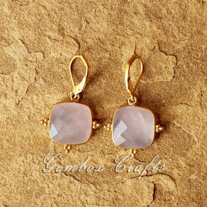 May include: A pair of gold earrings featuring a light pink square gemstone. The earrings have a simple design with a small gold loop at the top.