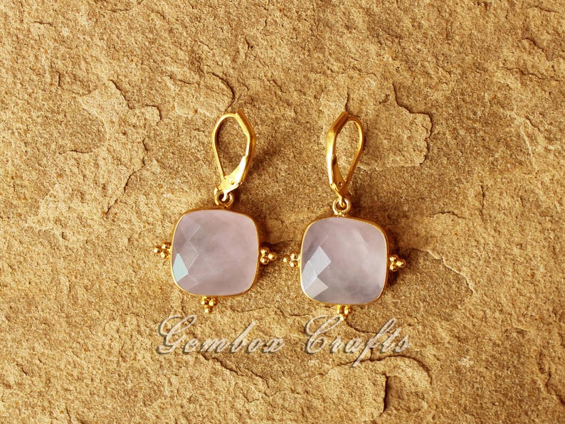 May include: A pair of gold earrings featuring a light pink square gemstone. The earrings have a simple design with a small gold loop at the top.