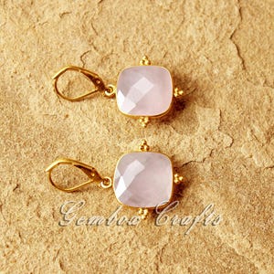 May include: A pair of gold earrings featuring pink square-shaped gemstones. The earrings have a delicate design with small gold beads around the edges of the gemstones.