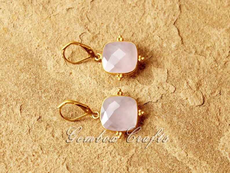 May include: A pair of gold earrings featuring pink square-shaped gemstones. The earrings have a delicate design with small gold beads around the edges of the gemstones.
