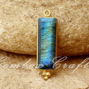 以下が含まれることがあります： 長方形の青いラブラドライトの宝石が付いたゴールドのペンダント。ペンダントの上部にベイルがあり、下部に小さなゴールドのボールが3つ付いています。