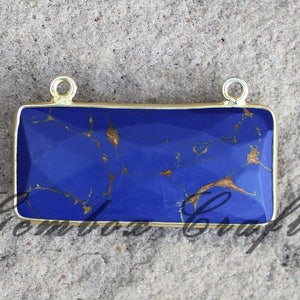 Puede incluir: Un colgante rectangular con una piedra preciosa azul y dorada engastada en un marco dorado. El colgante tiene dos bucles dorados en la parte superior para sujetar una cadena.