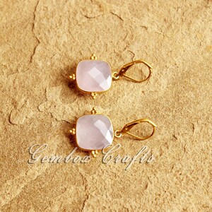 May include: A pair of gold earrings with pink gemstone drops. The earrings are square shaped and are accented with small gold beads.