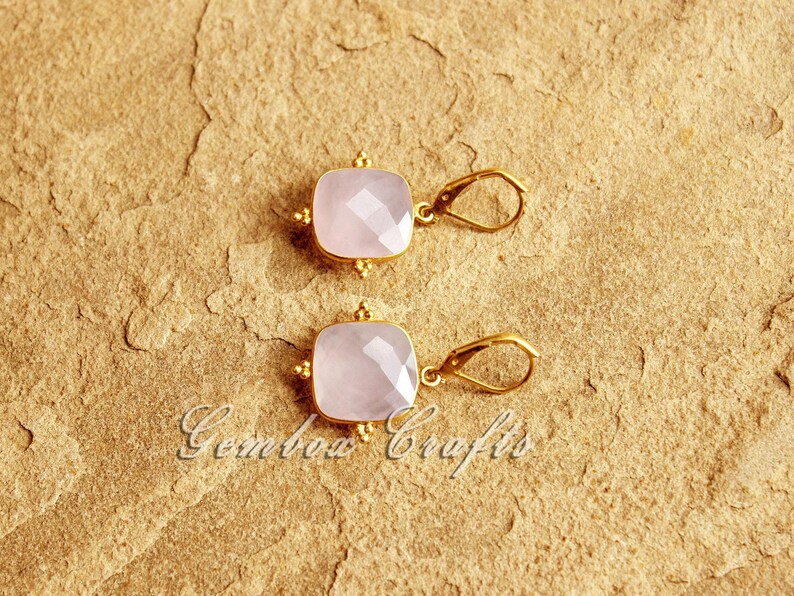 May include: A pair of gold earrings with pink gemstone drops. The earrings are square shaped and are accented with small gold beads.