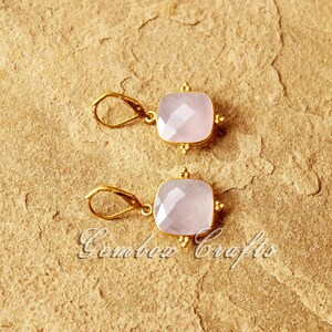 May include: A pair of gold earrings featuring a light pink square gemstone. The earrings have a delicate design with small gold beads surrounding the gemstone.