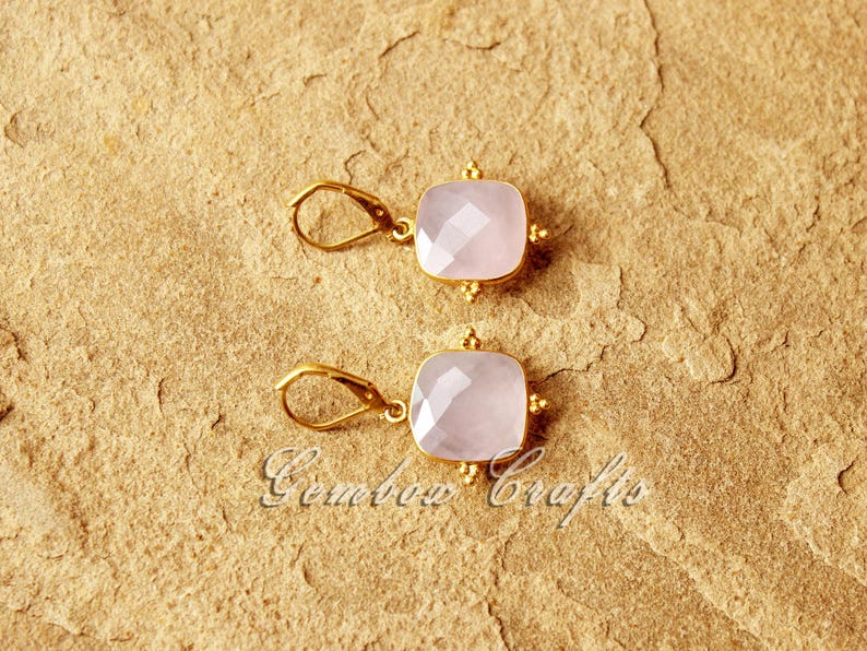 May include: A pair of gold earrings featuring a light pink square gemstone. The earrings have a delicate design with small gold beads surrounding the gemstone.