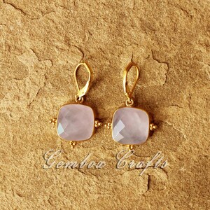 May include: A pair of gold earrings featuring a square-shaped pink gemstone. The earrings have a delicate design with small gold beads surrounding the gemstone.