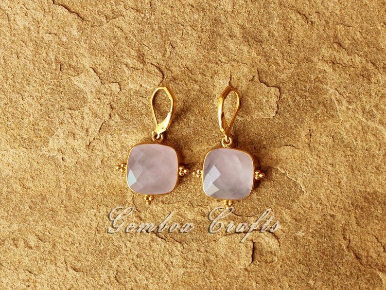 May include: A pair of gold earrings featuring a square-shaped pink gemstone. The earrings have a delicate design with small gold beads surrounding the gemstone.
