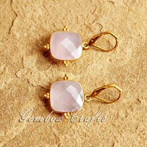 May include: A pair of gold earrings featuring pink square-shaped gemstones. The earrings have a simple design with a small gold loop for hanging.