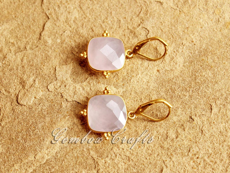 May include: A pair of gold earrings featuring pink square-shaped gemstones. The earrings have a simple design with a small gold loop for hanging.