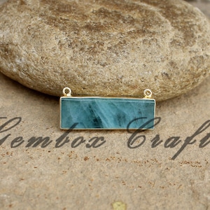May include: A rectangular pendant with a light blue gemstone set in gold metal. The pendant has two gold loops at the top for attaching a chain.