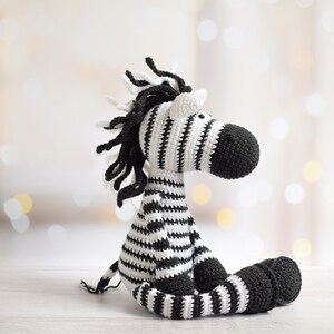 May include: A black and white striped crocheted zebra stuffed animal. The zebra is sitting with its legs crossed and has a long, black mane.