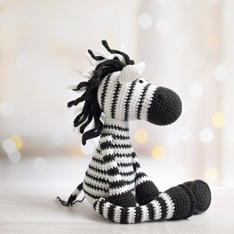 May include: A black and white striped crocheted zebra stuffed animal. The zebra is sitting with its legs crossed and has a long, black mane.