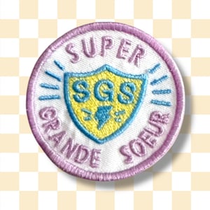 Peut inclure: Un écusson brodé blanc avec une bordure rose. L'écusson présente un bouclier bleu et jaune avec les lettres "SGS" en jaune. Le texte "SUPER GRANDE SOEUR" est écrit en cercle autour du bouclier.