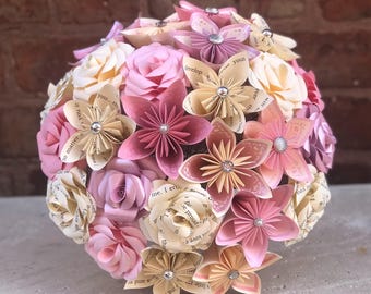 May include: A spherical bouquet of handcrafted paper flowers in shades of pink, cream, and beige. The arrangement features rolled paper roses and folded origami-style blossoms, many accented with small silver beads and printed text.