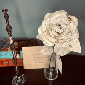 May include: A white paper rose made from the pages of a book, with a brown stem and a clear glass vase. The rose is sitting on a dark brown surface, next to a Harry Potter book and a wand.
