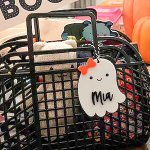 May include: Two black wire baskets filled with Halloween treats and toys. Each basket has a ghost-shaped tag with a name on it. Pink pennant flags read "Hey Boo" and "Trick or Treat". Orange pumpkins and Halloween decorations are in the background.