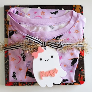 May include: A wrapped gift set featuring a lavender pajama top with a Halloween-themed print of bats, stars, and moons. The set is tied with a black and white striped ribbon and a gold tinsel garland. A white ghost-shaped tag with the name "Perse" is attached.