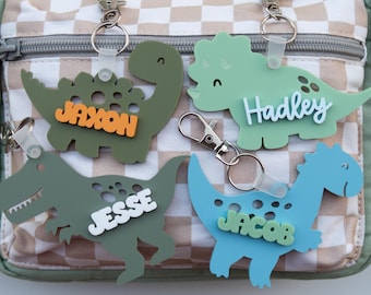May include: Four dinosaur-shaped keychains in green and blue, each with a different name: Jaxon, Hadley, Jesse, and Jacob. The keychains are attached to a bag with a checkered pattern.