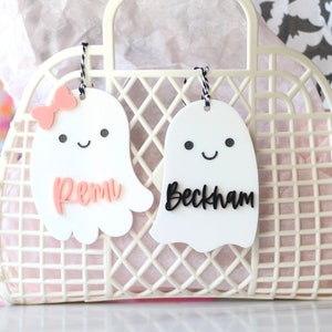May include: A white plastic tote bag with a grid pattern, holding pink tissue paper. Two white ghost-shaped tags hang from the handle, one with a pink bow and the name "Remi", the other with the name "Beckham" in black.