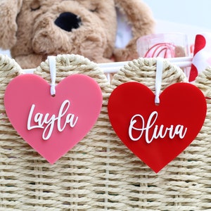 May include: Two heart-shaped gift tags, one pink and one red, with white script names "Layla" and "Olivia". The tags hang from a woven basket containing a stuffed animal and other gifts. The basket is made of light-colored woven material.