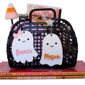 May include: A black plastic basket filled with Halloween-themed items, including a candy corn-shaped pen, a black cat charm, and two white ghost-shaped tags with names. The basket sits on top of a stack of children's books.