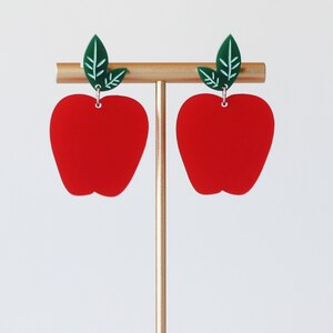 May include: A pair of red apple-shaped earrings with green leaf accents. The earrings are on a gold stand.