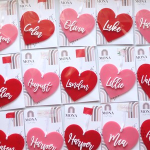 May include: A collection of heart-shaped ornaments in shades of pink and red, each with a name in white script. The ornaments are attached to cards with a rainbow design and the brand name "MONA".