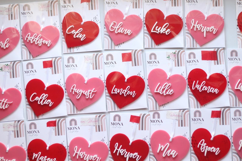 May include: A collection of heart-shaped ornaments in shades of pink and red, each with a name in white script. The ornaments are attached to cards with a rainbow design and the brand name "MONA".