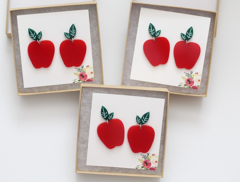 May include: Three small gift boxes each containing a pair of red apple earrings with green leaves. The earrings are made of polymer clay.