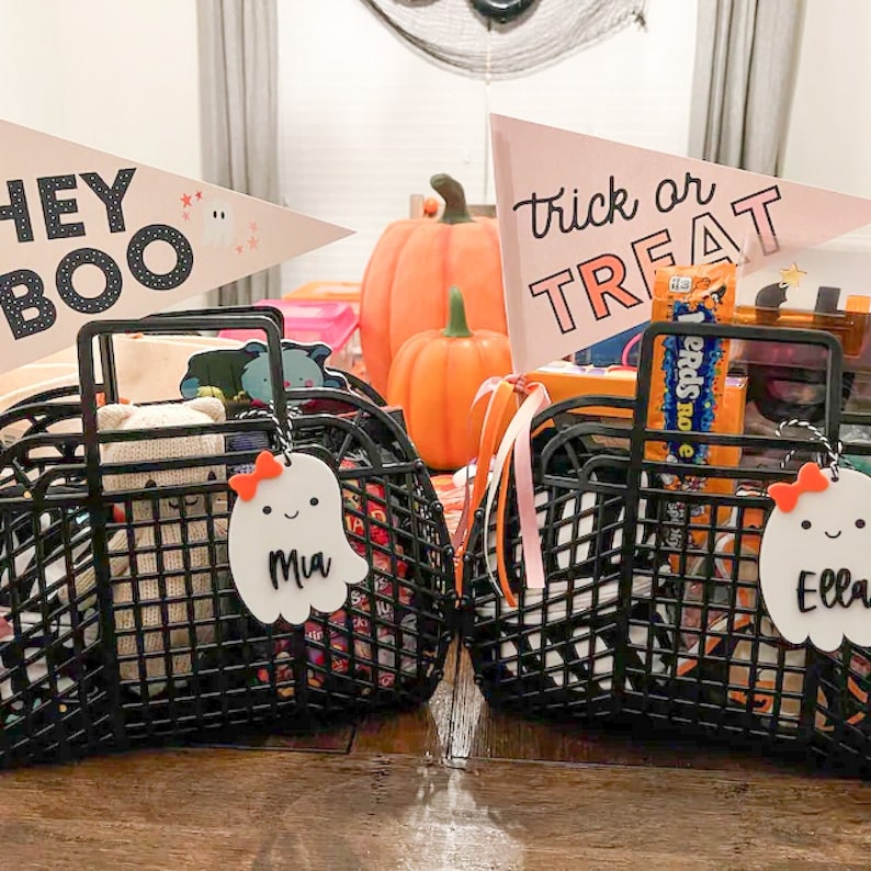 May include: Two black wire baskets filled with Halloween treats and toys. Each basket has a ghost-shaped tag with a name on it. Pink pennant flags read "Hey Boo" and "Trick or Treat". Orange pumpkins and Halloween decorations are in the background.