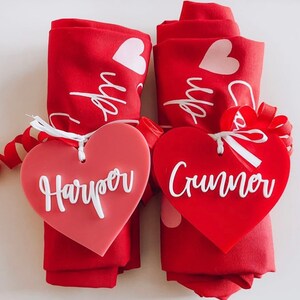May include: Two rolled red napkins with white heart designs and the word "up" printed on them. Each napkin is adorned with a heart-shaped tag. One tag is a gradient pink with the name "Harper" in white, and the other is red with the name "Crumner" in white.